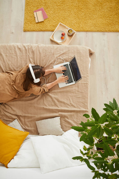 Top view of student girl in white headphones lying in bed and typing on laptop while searching for information on educational project - Фото, изображение