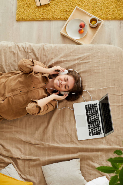 High angle view of happy girl with closed eyes lying on bed and listening to music in headphones - Foto, immagini