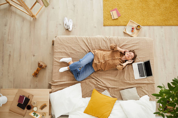Top view of jolly young woman in casual clothing lying on bed with laptop and listening to song in headphones - Foto, Imagen