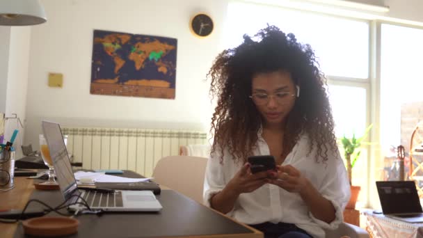 Mujer empresaria afroamericana seria con erabuds sentados en la mesa de la oficina en casa y leyendo mensajes en el teléfono celular durante el trabajo distante - Metraje, vídeo
