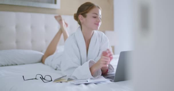 Full body of excited young female in white bathrobe lying on bed and stretching while watching movie on laptop during weekend at home - Footage, Video