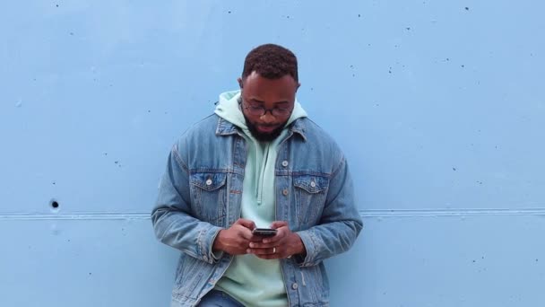 Dorosły hipster brodaty Afroamerykanin mężczyzna w luźnym stroju i okulary przeglądając smartfona i śmiejąc się stojąc przed beżową kamienną ścianą na ulicy - Materiał filmowy, wideo