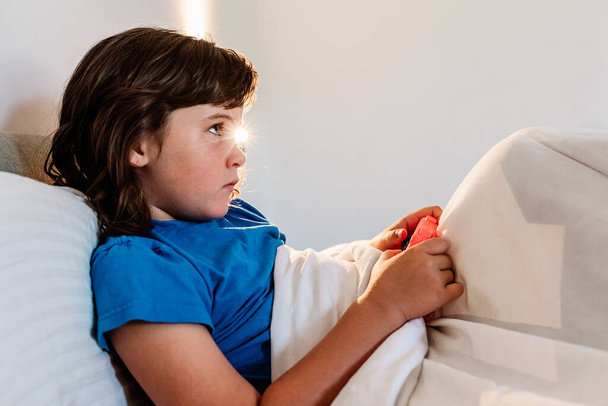 Vista lateral perturbada da menina com almofada de jogo jogando videogame na cama no quarto leve - Foto, Imagem