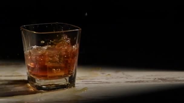 Persona vertiendo cubitos de hielo en vidrio con bourbon sobre mesa de madera en rayos sobre fondo negro - Imágenes, Vídeo