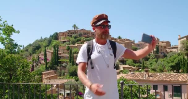 Vrolijke hipster bebaarde mannelijke toerist in stijlvolle zonnebril en pet selfie op smartphone tijdens een bezoek aan historische nederzetting tijdens de zomerreis - Video