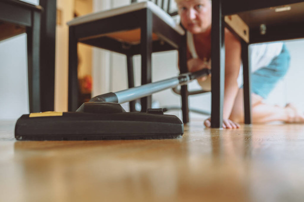 Женщина убирает квартиру с паркетным полом с пылесосом. женщина приседает с помощью пылесоса под обеденным столом дома. пылесос под - Фото, изображение