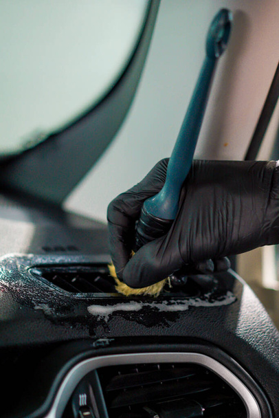 Eine Nahaufnahme eines Arbeiters in einer Autowaschanlage, der während des Detaillierungsprozesses das Armaturenbrett eines Luxusautos mit einer Bürste und Chemikalien säubert - Foto, Bild
