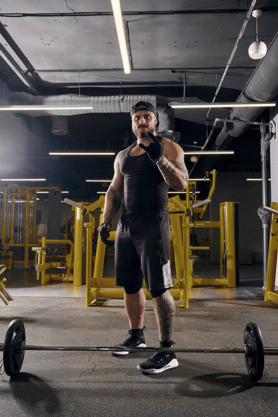 Attractive, tattooed, bearded man in black sport gloves, shorts, vest, cap and sneakers. Showing cool sign, barbell is on floor, going to do exercises for training his muscles. Dark gym. Full length - Photo, Image