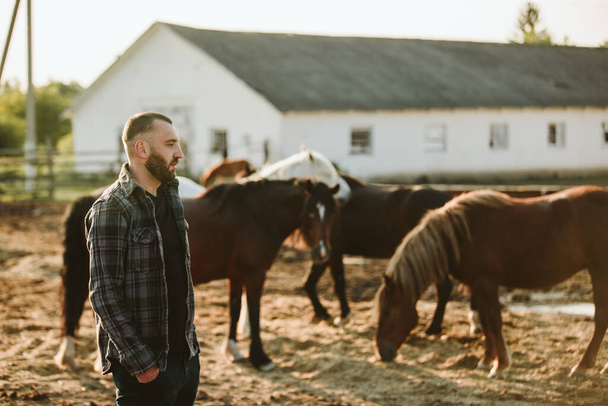 Portret van een jonge landarbeider, op de achtergrond van een hek met paarden. Een jonge boer zorgt voor paarden in de stallen. - Foto, afbeelding