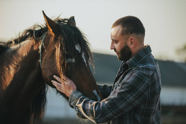 Un hombre acaricia un caballo. Un joven granjero barbudo se encarga de los caballos. - Foto, Imagen