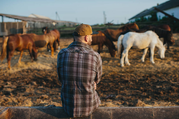 Retrato de un joven campesino, sobre el fondo de una cerca con caballos. Un joven agricultor se encarga de los caballos en los establos. - Foto, Imagen