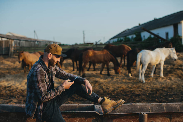 Молодой человек с телефоном в руках на заднем плане загона с лошадьми. Человек отдыхает после рабочего дня на ферме, на закате. - Фото, изображение