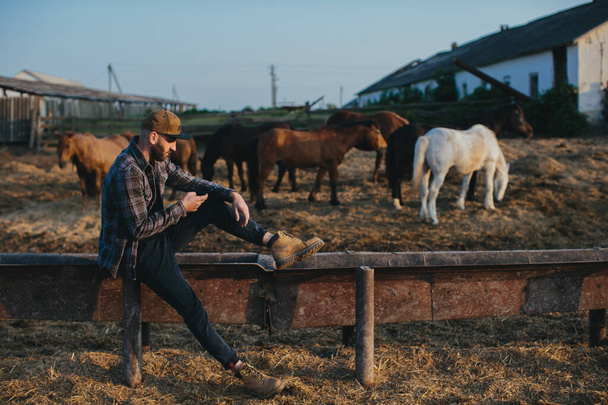 Un giovanotto con un telefono in mano sullo sfondo di un recinto con dei cavalli. Un uomo si riposa dopo una giornata di lavoro in fattoria, al tramonto. - Foto, immagini