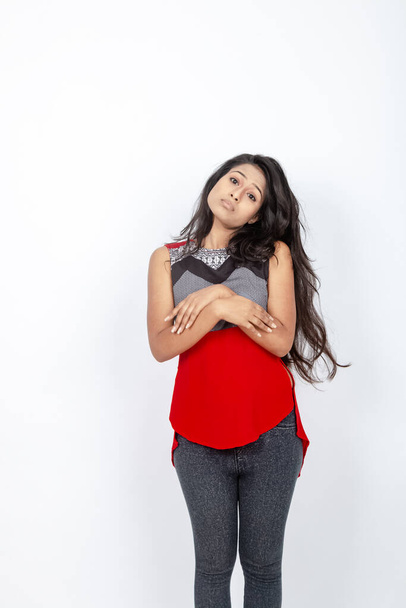 Studio shot of a young, beautiful Indian female model in casual wear wearing red and grey designer top and black jeans against white background. Female model. Fashion Portrait. - Photo, Image