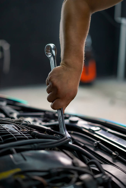 Nahaufnahme eines Autoservice-Mitarbeiters, der mit einem Schraubenschlüssel in der Hand ein teures Auto mit offener Motorhaube fixiert - Foto, Bild