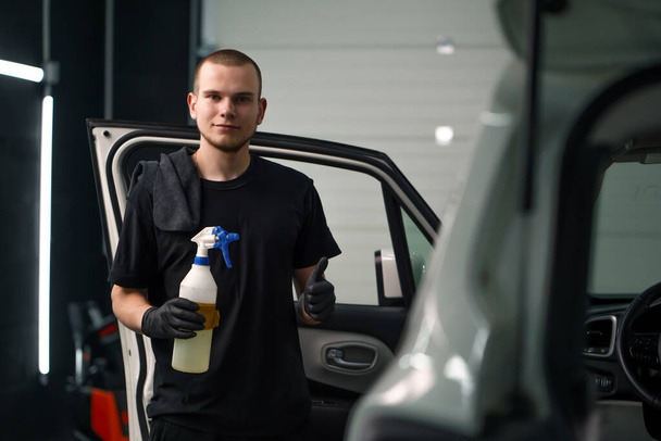 Retrato de un joven trabajador de lavado de coches sosteniendo una pistola de pulverización y mostrando un cartel de pulgares hacia arriba en el fondo de un SUV blanco en t proceso de desenredar - Foto, imagen