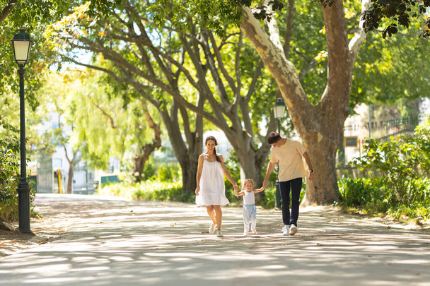 Маленькая девочка с родителями, гуляющими в парке, держась за руки. Средний выстрел - Фото, изображение