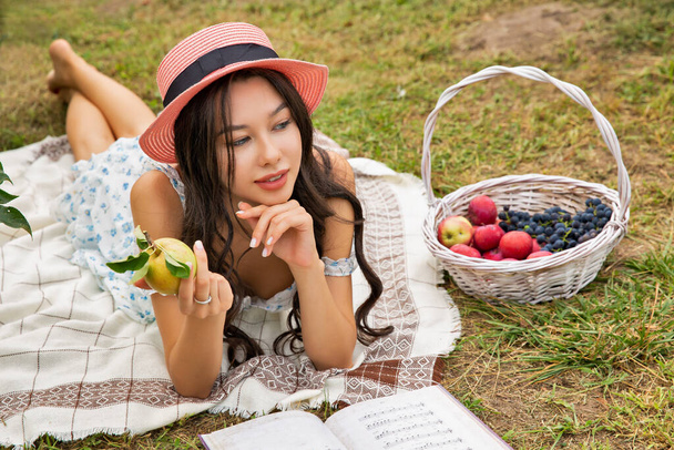 Όμορφη νεαρή μελαχρινή καυκάσια γυναίκα φορώντας καπέλο ξαπλωμένη στον κήπο και διαβάζοντας το βιβλίο. - Φωτογραφία, εικόνα