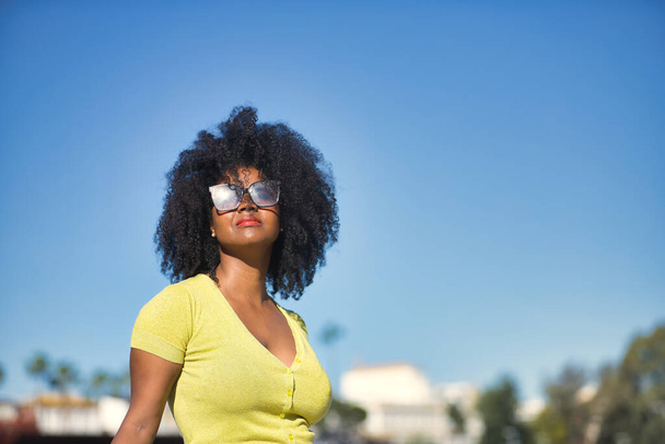 Πορτρέτο της νεαρής, όμορφης, μαύρης γυναίκας με αφρο μαλλιά, με κίτρινο t-shirt και γυαλιά ηλίου, κοιτάζοντας τον ήλιο, χαλαρή και ήρεμη. Concept διακοπές, ταξίδια, ήλιος, γυαλιά ηλίου, τρέχουσες, σύγχρονες. - Φωτογραφία, εικόνα