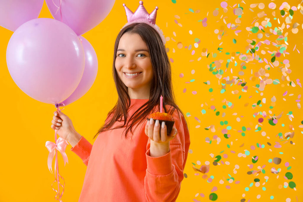 Mujer joven feliz con muffin de cumpleaños y globos sobre fondo amarillo - Foto, imagen