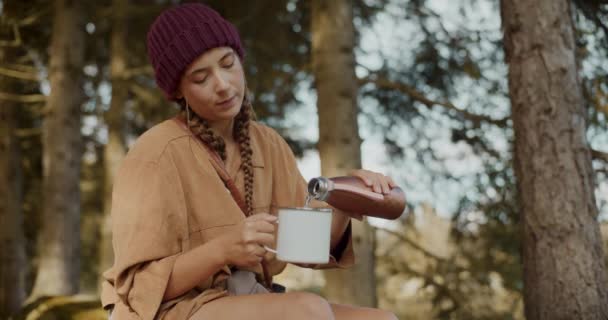 Jeune femme versant de l'eau dans une tasse tout en étant assis dans la forêt pendant les vacances d'été - Séquence, vidéo