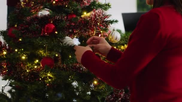 Close-up van medewerkers die kerstboom versieren met bloemenslingers, kerstverlichting en kerstballen op kantoor tijdens Secret Santa feest. Medewerkers die tijd samen doorbrengen op het werk, genieten van de feestelijke sfeer - Video