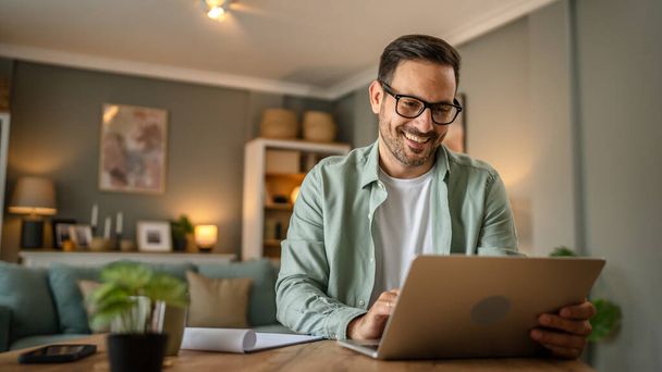 un homme adulte caucasien mâle avec barbe travail sur son ordinateur portable à la maison heureux sourire succès entrepreneur indépendant ou concept de travail à distance - Photo, image