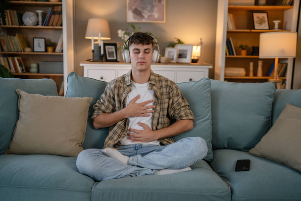 yksi teini mies nuori valkoihoinen mies käyttää kuulokkeita verkossa opastettu meditaatio käytännössä mindfulness jooga silmät kiinni kotona todellisia ihmisiä itsehoidon ilmentymä käsite kopioi tilaa sukupolven Z - Valokuva, kuva