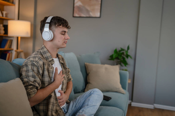 een tiener man jong Kaukasisch man gebruik hoofdtelefoon voor online geleide meditatie praktijk mindfulness yoga met ogen dicht thuis echte mensen zelfzorg manifestatie concept kopieer ruimte generatie Z - Foto, afbeelding