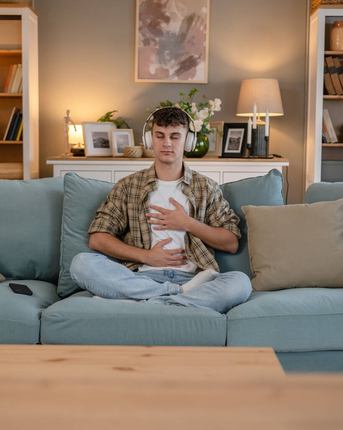 Ένας έφηβος άνδρας νεαρός καυκάσιος άντρας χρησιμοποιεί ακουστικά για online καθοδηγούμενη πρακτική διαλογισμού mindfulness yoga με τα μάτια κλειστά στο σπίτι πραγματικοί άνθρωποι αυτο-φροντίδα εκδήλωση έννοια αντίγραφο χώρο γενιά Z - Φωτογραφία, εικόνα