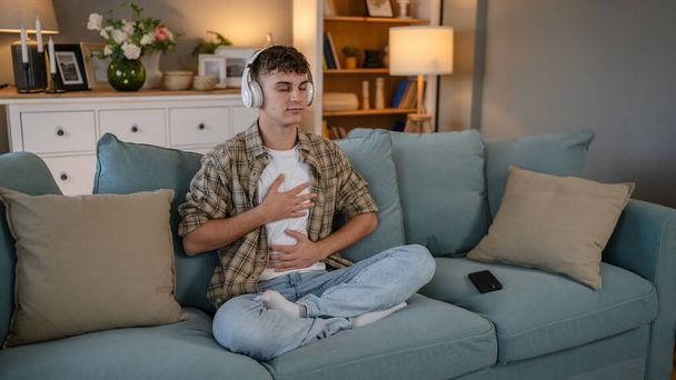 один подросток мужчина молодой кавказский мужчина использовать наушники для онлайн руководствоваться практикой медитации внимательность йога с закрытыми глазами на дому реальные люди самопроявление концепции самообслуживания копировать пространство поколения Z - Фото, изображение