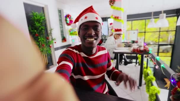 Αφροαμερικάνος εργάτης σε χριστουγεννιάτικο διακοσμημένο γραφείο φορώντας καπέλο Σάντα κινηματογραφώντας τον εαυτό του χρησιμοποιώντας selfie κάμερα, απολαμβάνοντας ωραία συνομιλία με τη φίλη πάνω από βίντεο στο διαδίκτυο κατά τη διάρκεια της χειμερινής περιόδου διακοπών - Πλάνα, βίντεο