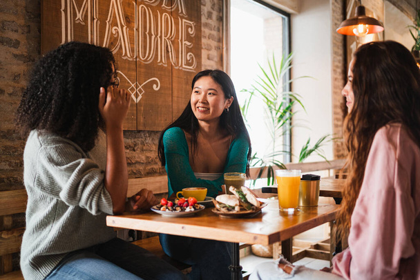 Τρεις χαμογελαστοί φίλοι κουτσομπολεύουν ενώ τρώνε δεκατιανό σε καφετέρια. - Φωτογραφία, εικόνα