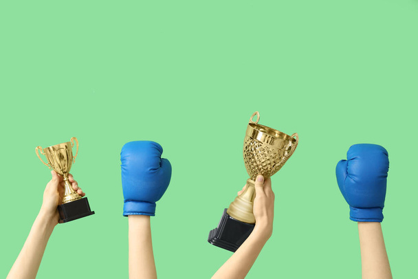 Manos femeninas con copas doradas y guantes de boxeo sobre fondo verde - Foto, Imagen