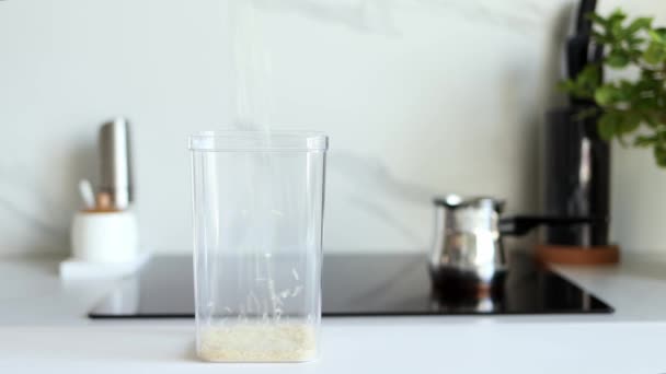 El arroz crudo blanco se vierte en un recipiente transparente sobre el fondo de una cocina moderna. Concepto de alimentación y nutrición saludables. Productos seguros, ecológicos y saludables. Envases ecológicos, plagas - Metraje, vídeo