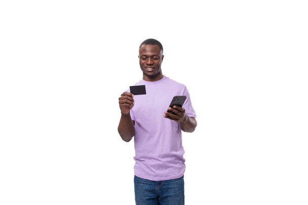 jovem e bonito homem africano em uma camiseta lilás usa um smartphone e um cartão de crédito em um fundo branco com espaço de cópia. conceito de publicidade. - Foto, Imagem