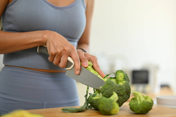 Mujer joven cortando brócoli fresco en la tabla de cortar, preparando comida saludable en la cocina. - Foto, imagen