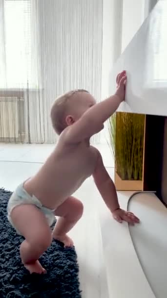 small child in a diaper stands on his feet near the white fireplace. Trying to get something with his hands - Footage, Video