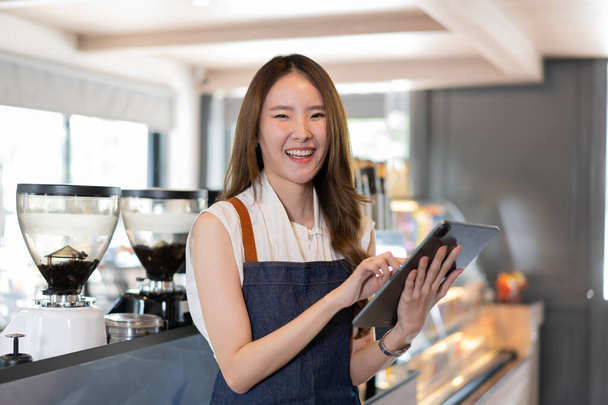 Young Asian female coffee shop owner and barista using a digital tablet and looking at a camera. Millennial Asia woman smiling and working at cafe and restaurant indoors. Technology usage  - Foto, Bild