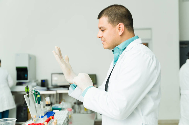 Hispanic professional chemist with a white lab coat putting on gloves working in a medical laboratory doing blood medical tests - Photo, Image