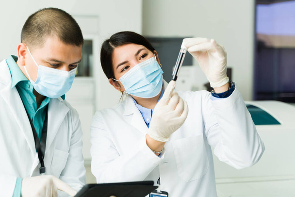 Kaukasische chemici met een gezichtsmasker en labjas met een bloedmonster in een reageerbuis terwijl ze medische tests in het laboratorium doen - Foto, afbeelding