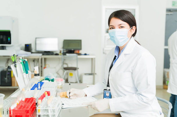 Piękna szczęśliwa kobieta z maską na twarzy uśmiechnięta pracująca jako chemik lub technik laboratoryjny pracująca nad wynikami badań próbek krwi  - Zdjęcie, obraz