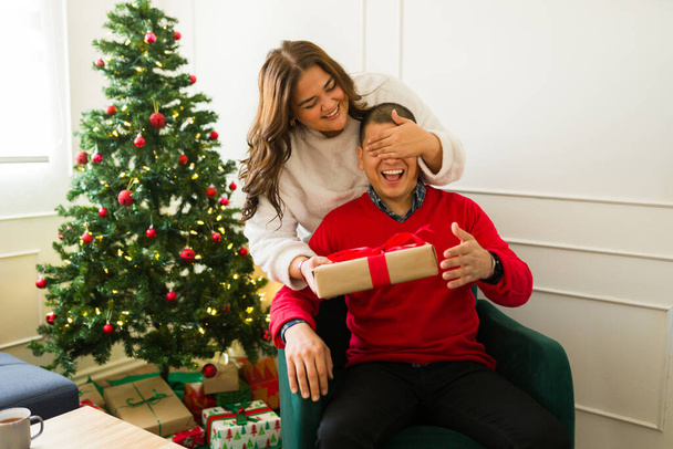 Ενθουσιασμένη γυναίκα συν μέγεθος καλύπτει τα μάτια του φίλου της και δίνει ένα όμορφο δώρο έκπληξη στο σύζυγό της για τα Χριστούγεννα  - Φωτογραφία, εικόνα