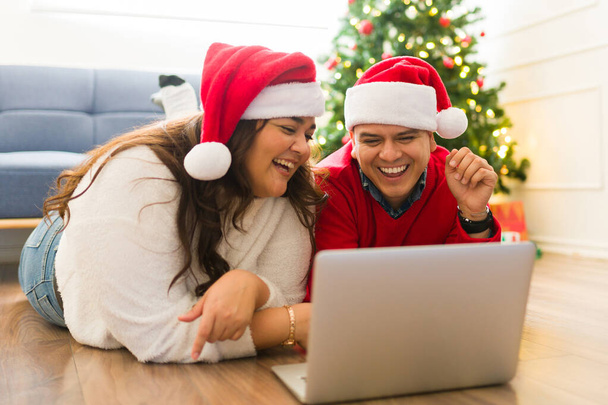 Couple latin excité rire et s'amuser tout en faisant du shopping en ligne sur l'ordinateur portable acheter des cadeaux de Noël pour la famille - Photo, image