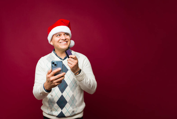 Glücklicher hispanischer Mann beim Online-Shopping auf seinem Handy und beim Bezahlen mit Kreditkarte für seine Geschenke während der Weihnachtsfeiertage - Foto, Bild