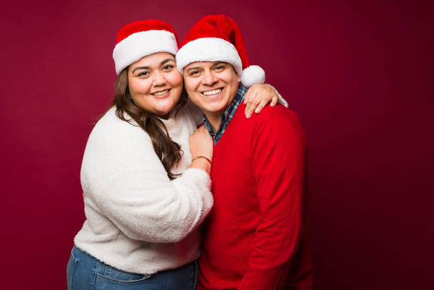 Hermosa pareja mexicana feliz abrazándose usando sombreros de santa juntos sonriendo mientras celebran las vacaciones de Navidad - Foto, Imagen