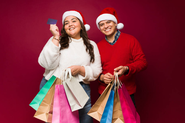Cheerful fat woman with an hispanic man paying with a credit card for their christmas presents after going shopping together - 写真・画像