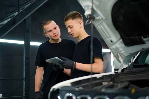 İki oto tamircisi kaputu açık beyaz SUV 'nin yanında duruyor ve arabayı tamir ederken ellerinde tuttukları tablete bakın. - Fotoğraf, Görsel