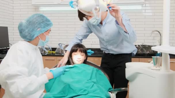 Aasian naispuolinen pediatrinen hammaslääkäri tarkistaa ja tutkii tytön hampaat isänsä kanssa, jota kannustetaan hammasklinikalla, hyvinvointihygieniassa ja ammatillisessa oikomishoidossa lastensairaalassa.. - Materiaali, video