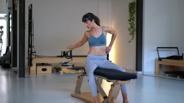 Soustředěná fit samice sedí na lavičce a protahuje paže na pilates stroje během tréninku - Záběry, video